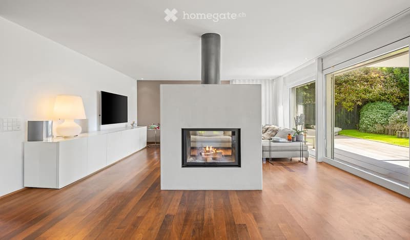 Modern living room with a central fireplace, large windows offering a view of the garden, a sofa, coffee table, floor lamp, and television.