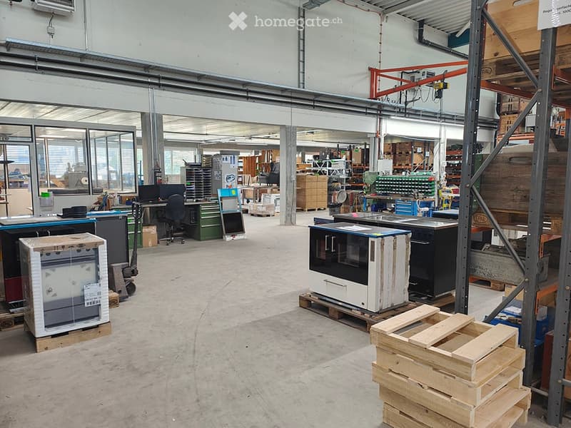 A spacious industrial workshop with large windows, metal shelving, workbenches, and various equipment including a refrigerator, oven, and storage boxes.