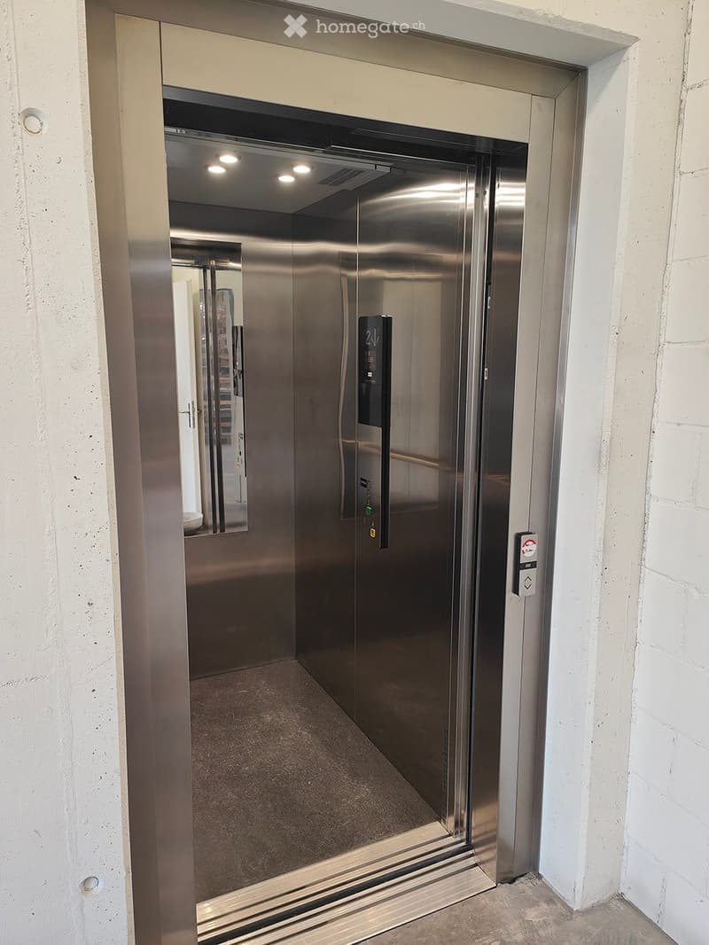 Modern hallway with a stainless steel elevator door, concrete walls, and a carpeted floor.