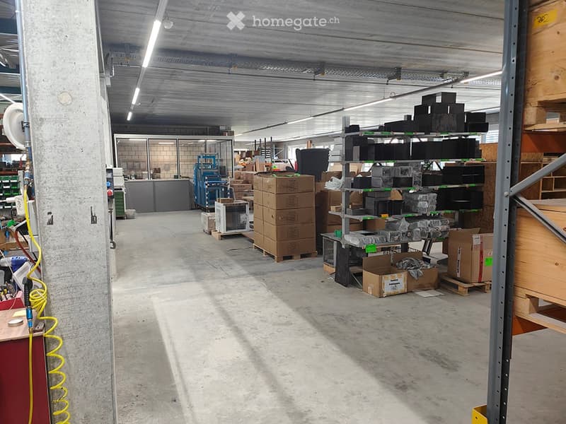 A spacious industrial-style warehouse with exposed metal beams and concrete floors. Shelves and metal racks hold various items, including cardboard boxes and equipment.