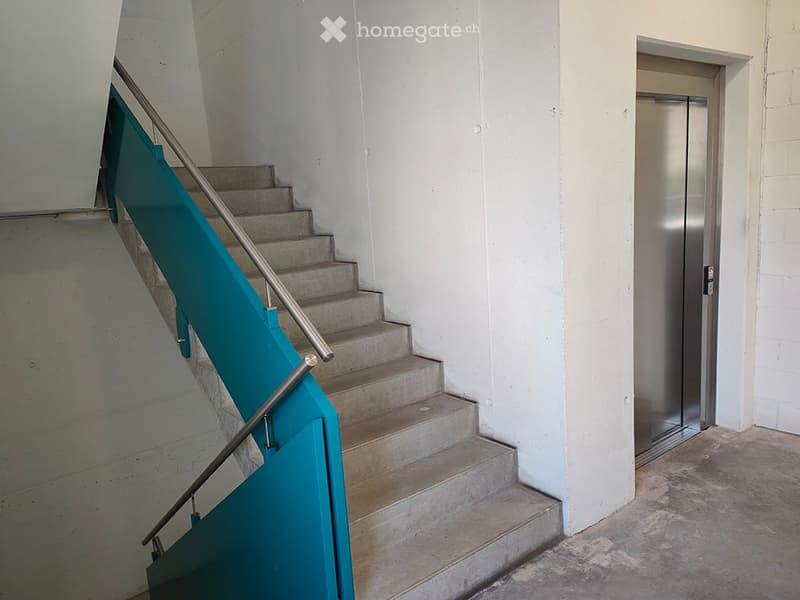 Modern hallway with a staircase featuring concrete steps and a teal handrail, alongside an elevator door.