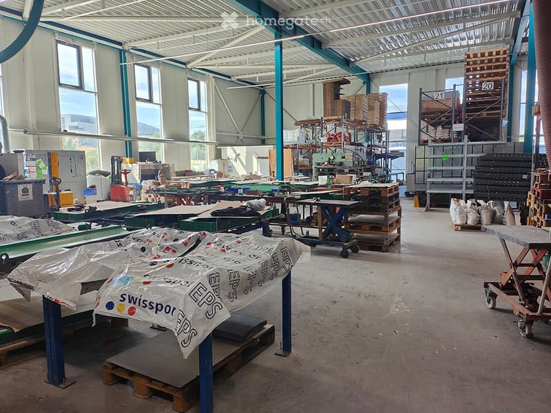 Industrial workshop with workbenches, pallets, shelves, machinery, and storage bins. Large windows provide natural light, and the space is organized for manufacturing or assembly.
