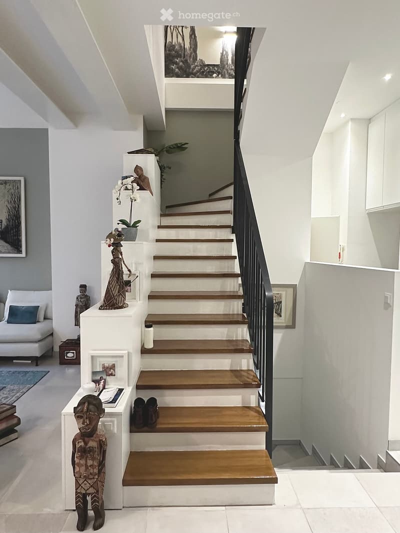 Modern hallway with a staircase featuring wooden steps and white risers, black metal railing, decorative items including sculptures, framed pictures, and a potted plant on a white console table. The hallway has white walls, a sleek design, and a view into