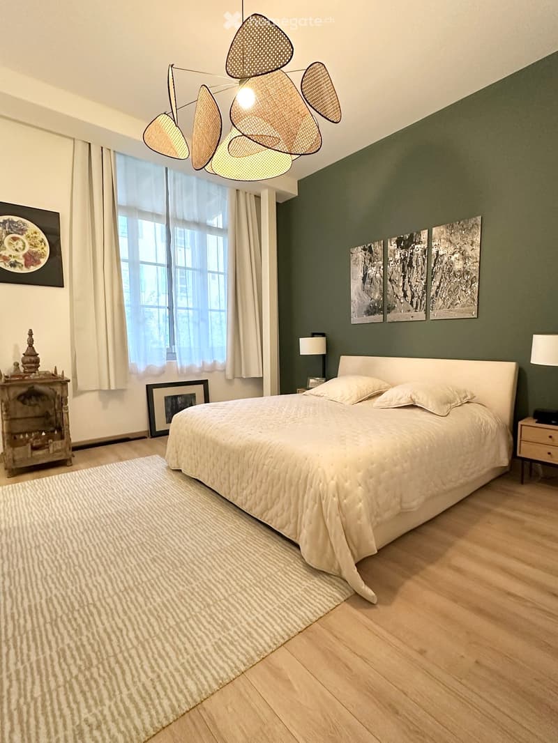 Modern bedroom with a large bed, white bedding, wooden nightstands, lamps, and artwork on the walls. Features large windows with curtains, a fireplace, and a decorative wooden cabinet.