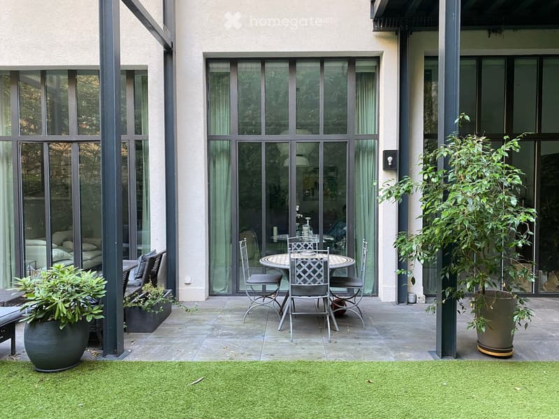 Modern outdoor patio with a round table, chairs, a bench, and potted plants, connected to a living space through large glass doors.