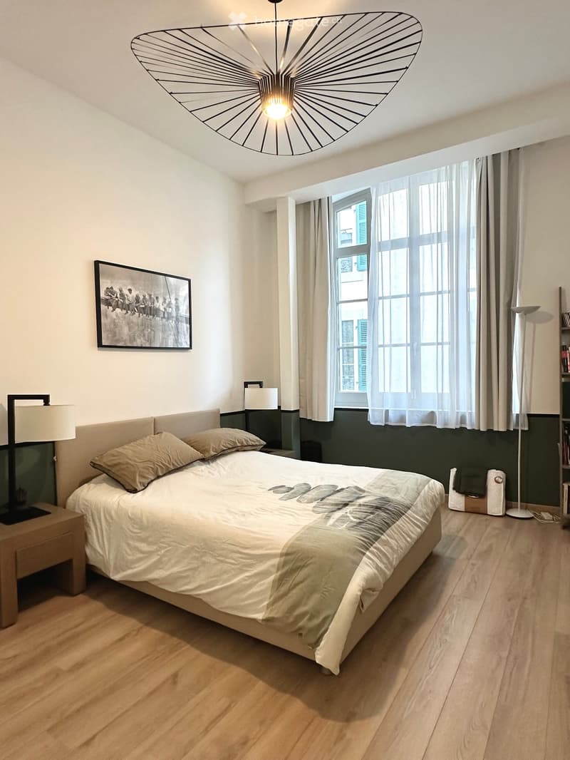 Modern bedroom with a large bed, nightstand, lamps, and a large window with curtains.