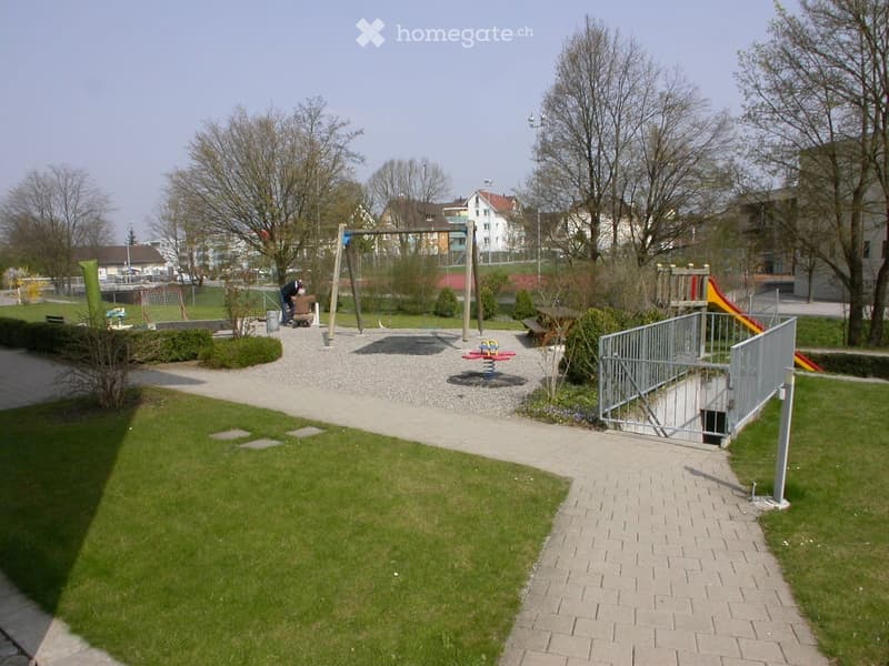 A well-maintained park area with a playground, swings, a slide, a small grassy lawn, paved walkways, and surrounding trees.