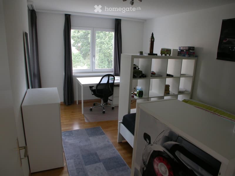 A furnished bedroom with a bed, desk, chair, shelf, and rug. The room has wooden flooring and large windows with curtains.