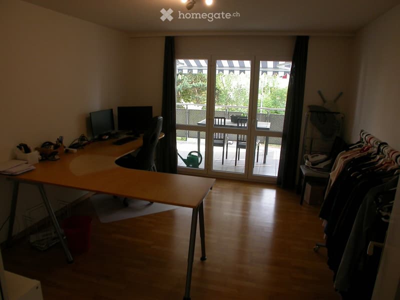 A furnished room with a wooden desk, computer, chair, and a clothing rack. Large windows and a balcony are visible.
