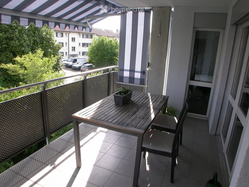 Balcony with a wooden table, two chairs, and a potted plant, overlooking a green area with parked cars.