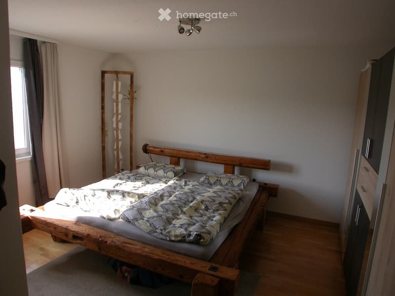 Cozy bedroom with wooden bed, patterned bedding, nightstand, and large mirror. Light-colored walls and wooden flooring.