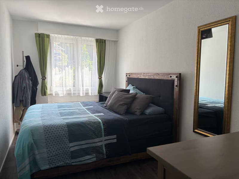 Cozy bedroom with a wooden bed, dark headboard, blue and grey bedding, a large mirror, and green curtains on the window.