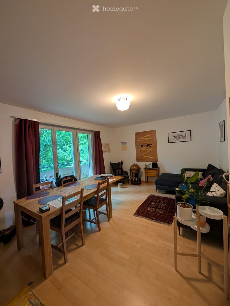 Bright living and dining area with wooden flooring, large sliding glass doors leading to a balcony, a dark-colored sofa, a wooden dining table with chairs, side tables, and decorative items including a globe and artwork.