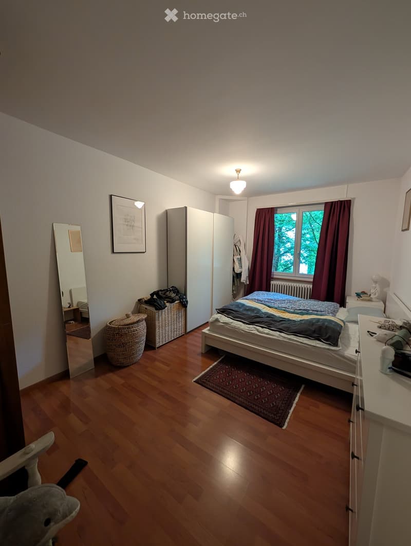 Cozy bedroom with wooden flooring, a single bed with colorful bedding, a wardrobe, a mirror, a nightstand, and a wicker basket. The room features large windows with dark curtains and a small rug on the floor.