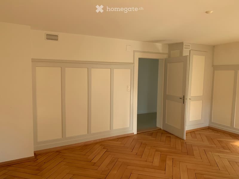 Empty room with wooden herringbone flooring, white walls, and multiple doors leading to other spaces.
