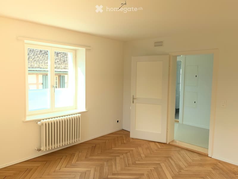 Empty room with light-colored walls, herringbone wooden flooring, a window with a radiator below, and an open door leading to another room.
