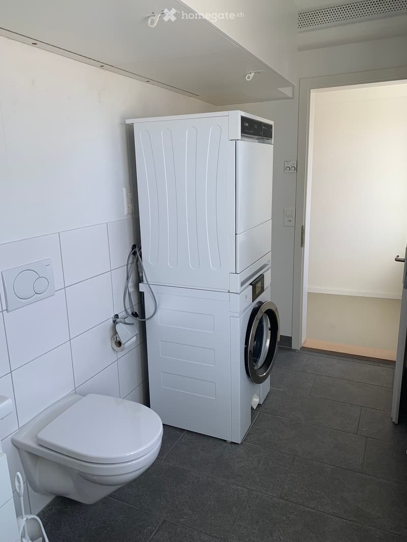 Modern bathroom with a toilet, washing machine, and dryer. The space features white tiled walls, a dark tiled floor, and a door leading to another area.