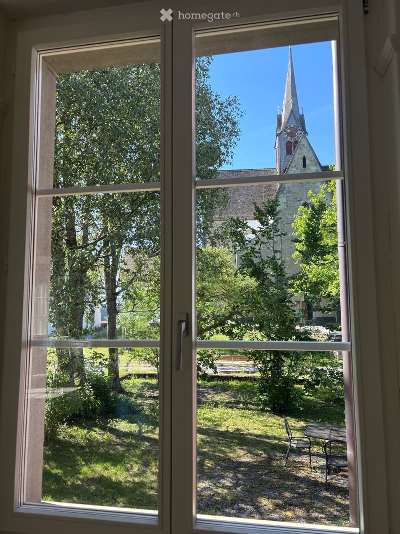View from a window showing a garden area with a table and chair, surrounded by trees and a church with a tall spire in the background.