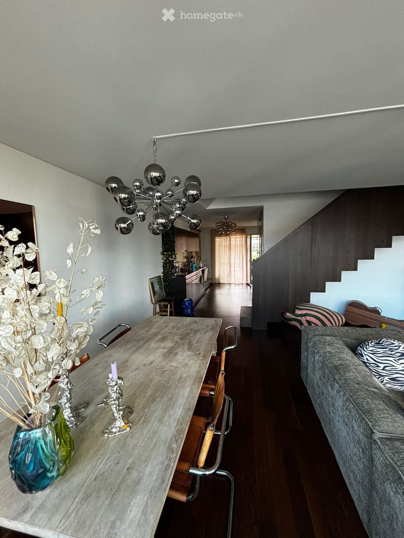 Modern living room with a grey sofa, wooden dining table, chairs, decorative vase with dried flowers, candle, and a chandelier. The room features a staircase leading to another level and a view into a kitchen area with cabinets and appliances.