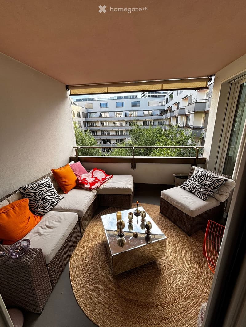 Cozy balcony with a corner sofa, coffee table, and chair, decorated with colorful cushions and a patterned throw, overlooking a cityscape with buildings and greenery.
