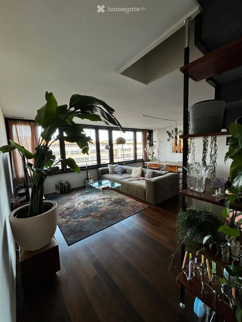 Modern living room with large windows, wooden flooring, a grey sofa, coffee table, dining area with table and chairs, decorative plants, and candle holders.