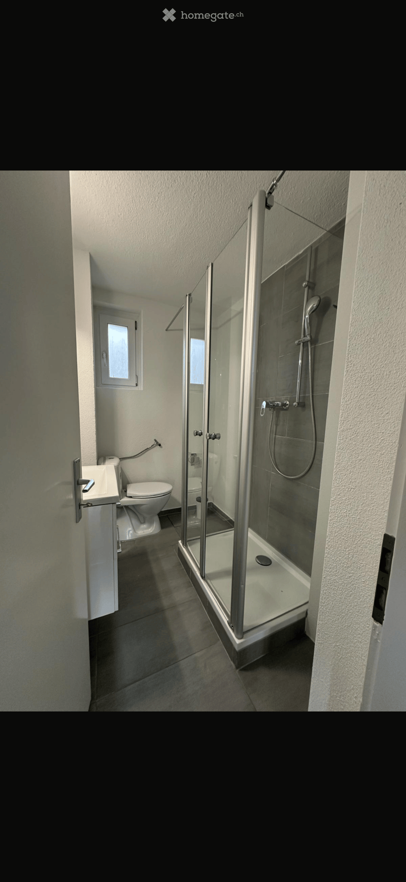 Modern bathroom with a glass-enclosed shower, toilet, and sink. The space features tiled walls and a small window for natural light.