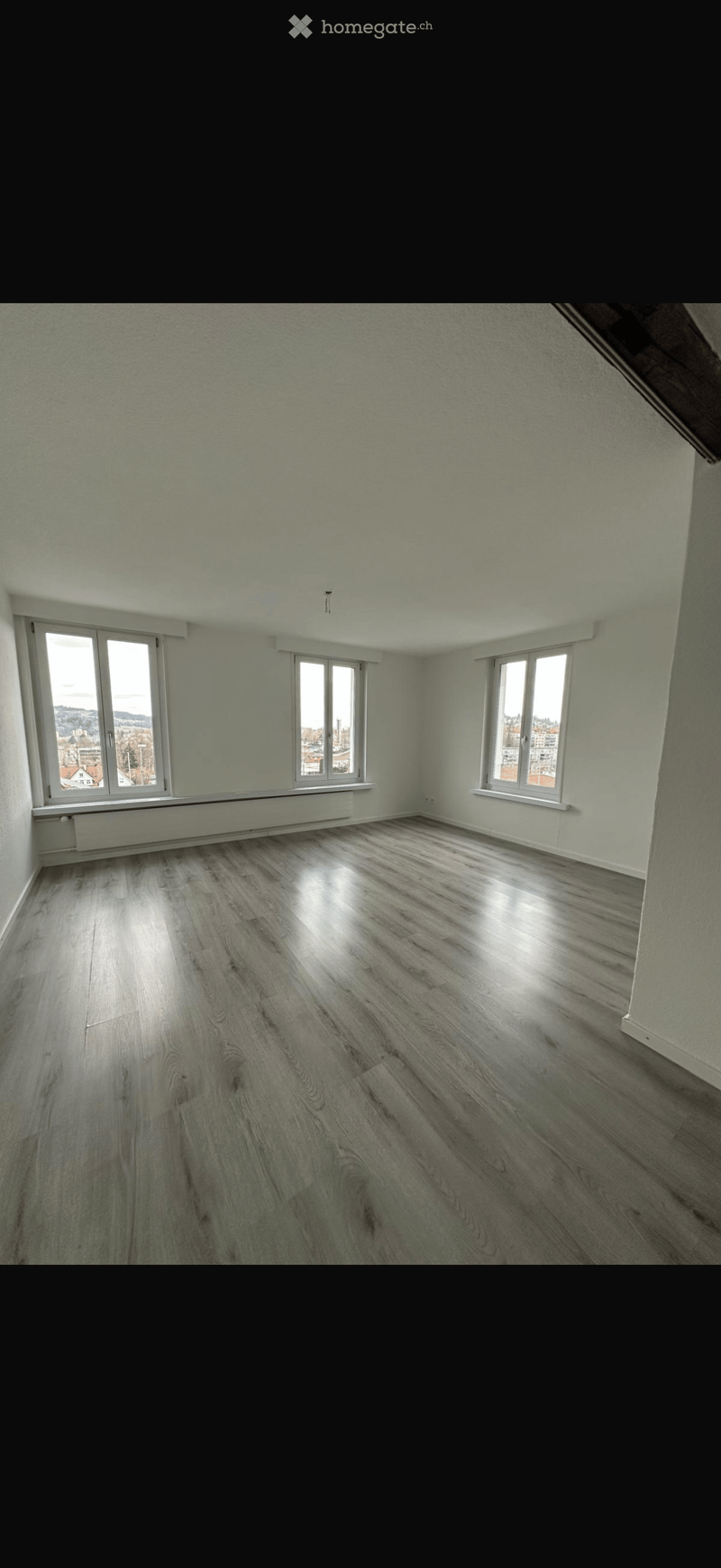 Empty room with light-colored walls, wooden flooring, and multiple windows offering a view of the outside.