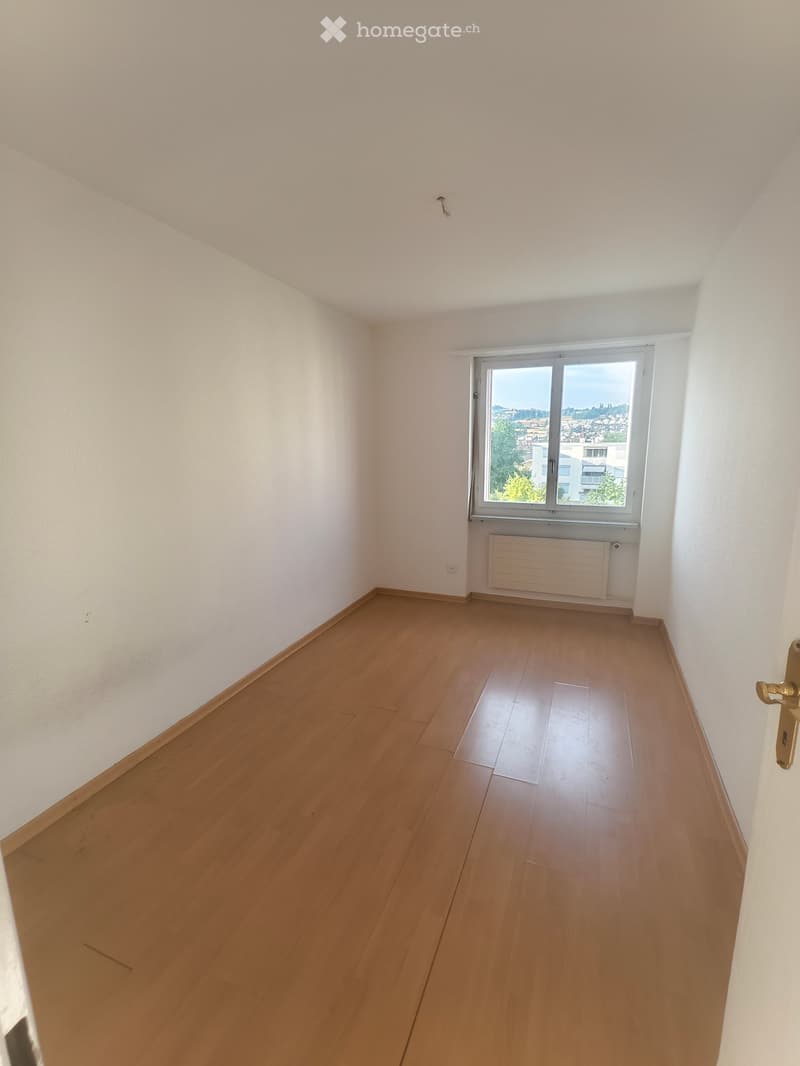 Empty room with wooden flooring, white walls, and a large window offering a city view. No furniture or equipment is present.