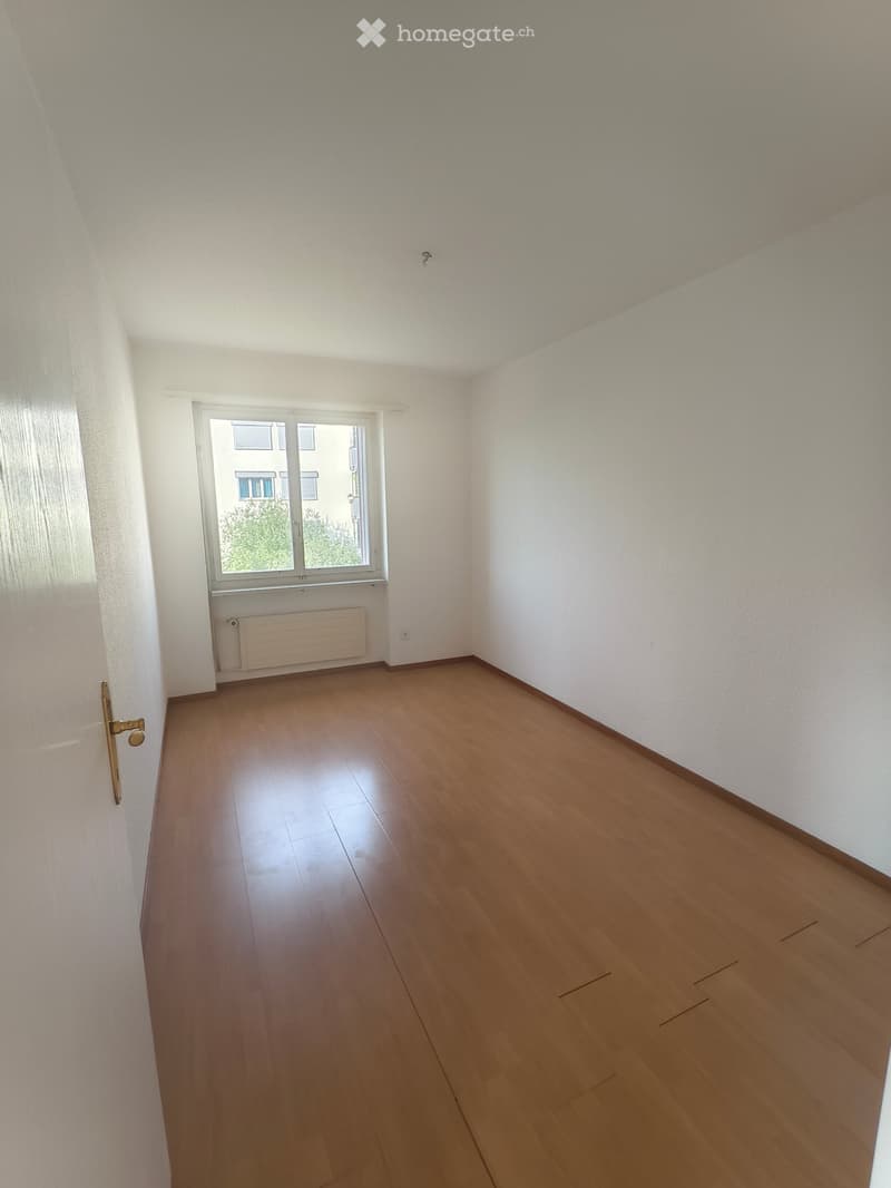 Empty room with wooden flooring, white walls, and a window with a radiator below it. The room has a door with a brass handle on the left side.