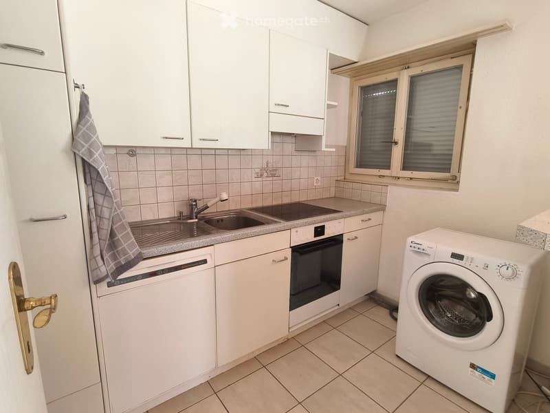 Modern kitchen with white cabinets, granite countertop, sink, dishwasher, oven, and washing machine. Features a window with blinds and tiled backsplash.