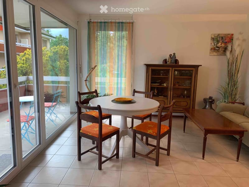 Bright living and dining area with large windows and balcony access, featuring a sofa, coffee table, dining table with chairs, and a wooden cabinet.