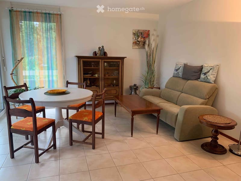 A well-furnished living and dining area with a beige sofa, wooden coffee table, round dining table with four chairs, a wooden cabinet, and decorative items including a plant and artwork.