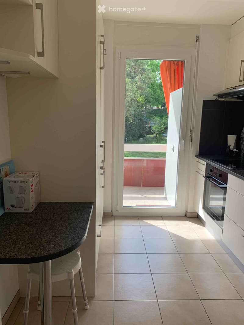 Modern kitchen with white cabinets, tiled floor, and a black countertop. A white stool is placed near the counter, and an electric kettle box is visible on the counter. An open sliding door leads to a balcony with a view of greenery.