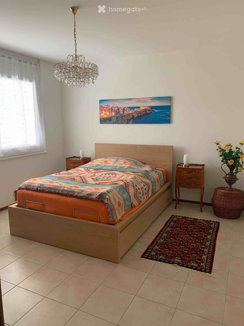 Cozy bedroom with a wooden bed, colorful bedspread, nightstands, chandelier, painting, and a wicker basket with flowers.
