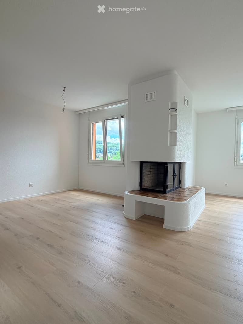 Empty room with light wooden flooring, white walls, and a built-in fireplace with a wooden surround. Large windows offer a scenic view.