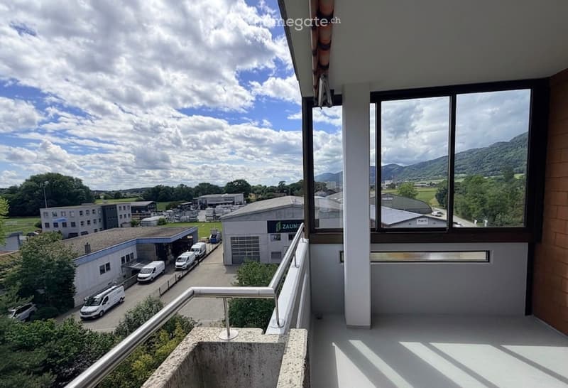 Balcony with large windows offering a view of an industrial area with buildings, vans, and surrounding landscape.