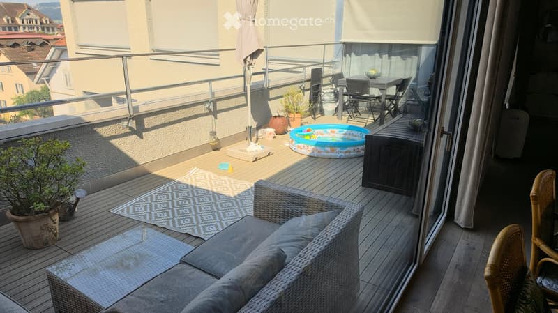 Balcony with wooden decking, grey wicker sofa, small table, chairs, potted plants, and a small inflatable pool.