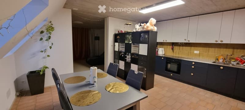 A modern kitchen and dining area with a grey dining table, four chairs, and a dark blue cabinetry. The kitchen features a sink, oven, and various appliances, with a small plant in a pot near the window.