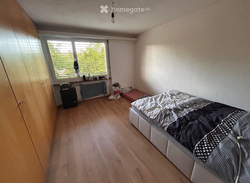Bedroom with wooden flooring, a bed with grey and black bedding, a wardrobe, a radiator, and a window with blinds.