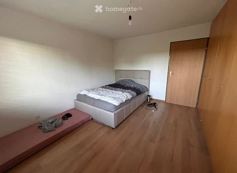 Bedroom with a grey upholstered bed, a matching nightstand, and a small bench at the foot of the bed. Wooden flooring and white walls.