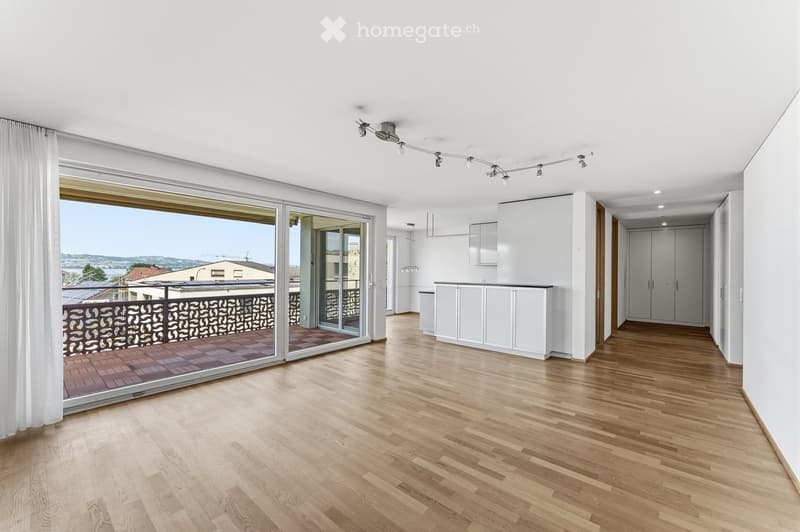 Bright, empty living space with wooden flooring, large sliding glass doors leading to a balcony, white cabinetry, and modern track lighting.