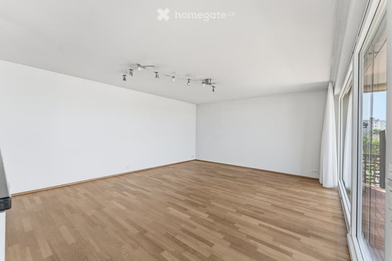 Empty room with white walls, wooden floor, and large windows with white curtains. Track lighting is mounted on the ceiling.