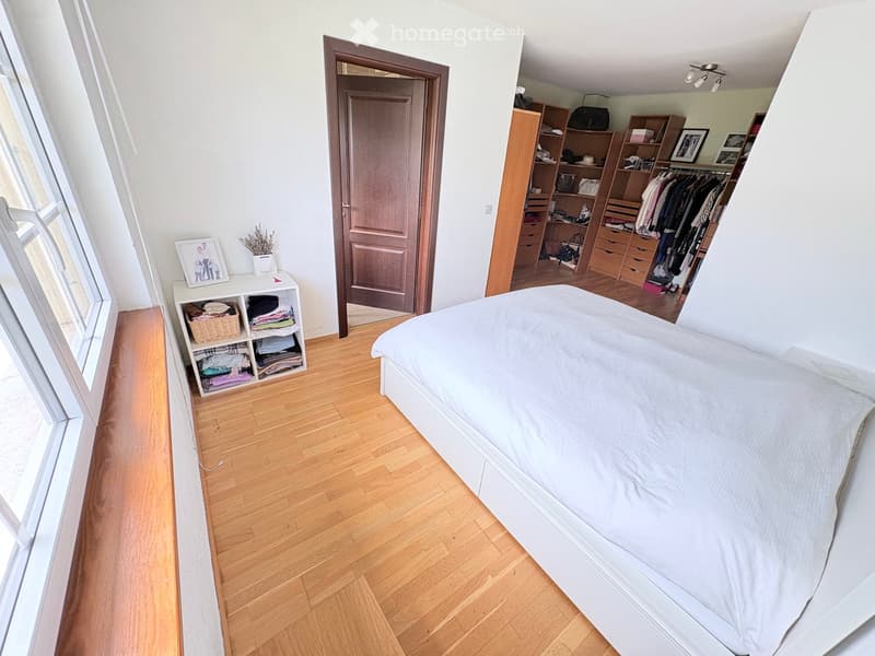 Bright bedroom with wooden flooring, a neatly made bed, a built-in wardrobe, and a small storage unit with folded clothes and a basket.