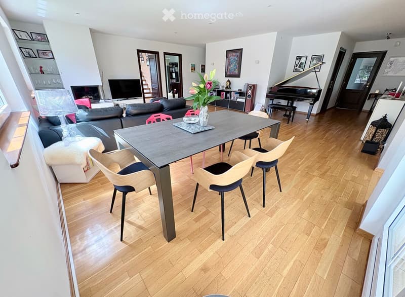 Modern living room with hardwood flooring, a black leather sofa, a grey dining table with four chairs, a grand piano, a television, and a fireplace. The room features a flower vase on the table and a bookshelf in the background.