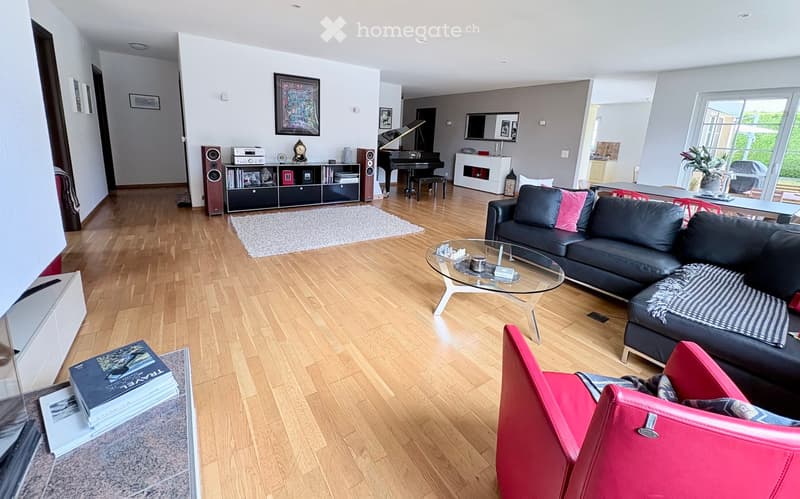Modern living room with wooden flooring, a black sectional sofa, a glass coffee table, a red leather armchair, a bookshelf, a piano, and a fireplace.
