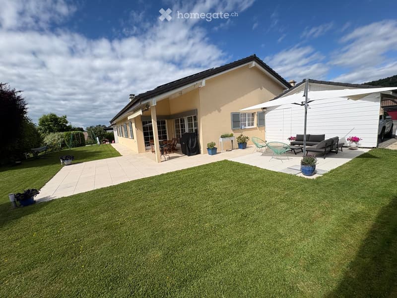 A well-maintained garden with a lawn, patio seating area, outdoor furniture, and a covered patio connected to a house.