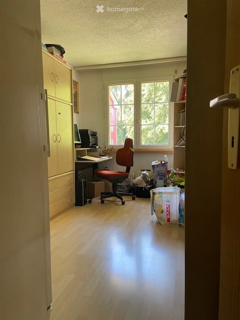 A furnished room with a wooden floor, a desk with a computer, a red office chair, a cabinet, and a bookshelf with books and other items.