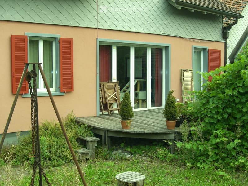 Garden area with a wooden deck, potted plants, wooden chairs, and a small bench. The house has a light-colored exterior with red shutters and a raised ground floor.