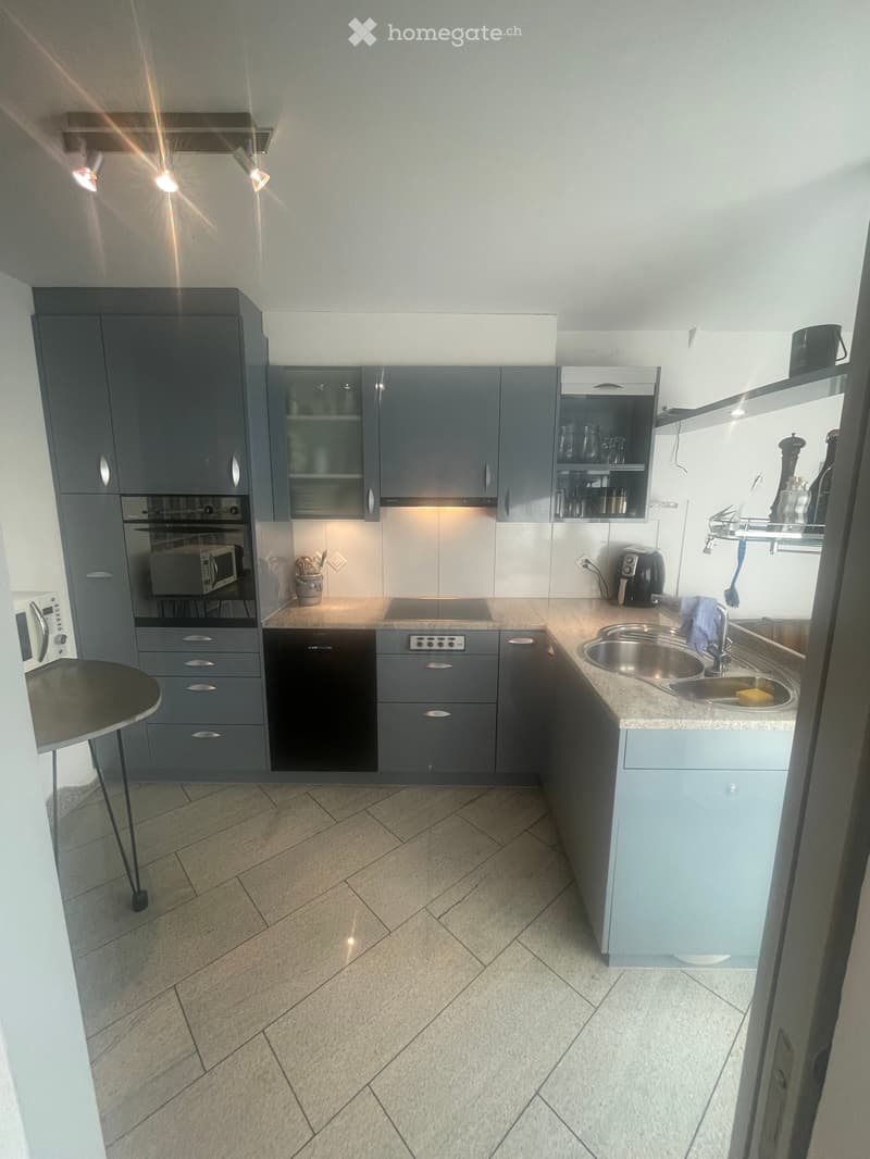 Modern kitchen with grey cabinets, a central island, and stainless steel appliances including a sink, microwave, and dishwasher. The countertop has a marble finish, and the floor is tiled.