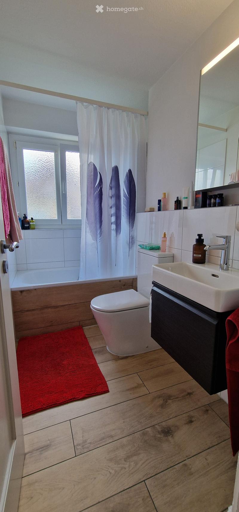 Modern bathroom with wooden flooring, white tiled bathtub, sink with modern faucet, toilet, and a large mirror above the sink. The space includes a red bath mat, a pink towel, and various toiletries on the sink counter.